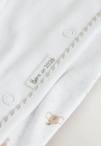 Witte katoenen stof met kleine dierenpatronen in beige en bruin. Opvallende details zijn drukknopen en een label met de tekst "Geboren in 2026."