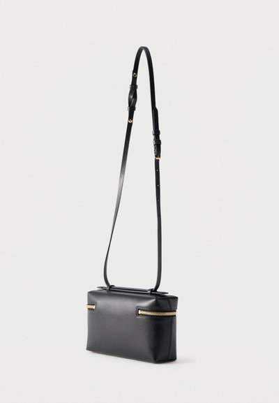 Μαύρη ορθογώνια δερμάτινη τσάντα crossbody με χρυσά φερμουάρ στα πλευρά και ρυθμιζόμενο λεπτό ιμάντα ώμου, πάνω σε απλό φόντο.