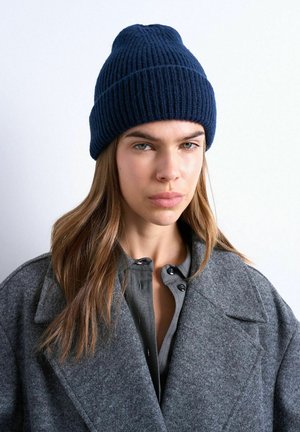 Woman with long brown hair wearing a navy knit beanie, gray buttoned shirt, and oversized gray wool coat against a plain white background.