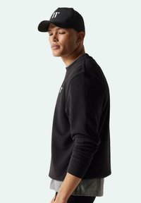 Sudadera negra con cuello redondo y puños ajustados, superpuesta sobre una camiseta gris clara. Gorra negra con logo en blanco, vista de lado.