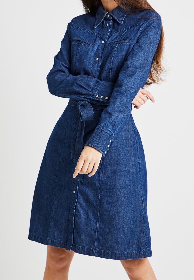 Femme portant une robe en denim bleu foncé longueur genou avec manches longues, devant boutonné, poches poitrine et ceinture nouée à la taille.