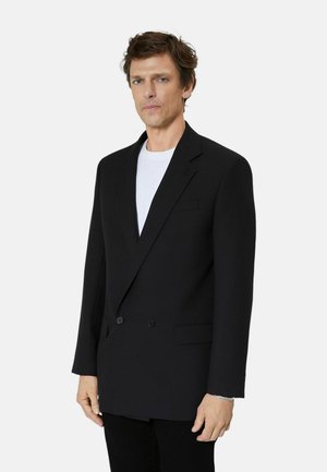 Mand iført en sort dobbeltradet blazer over en hvid skjorte, stående mod en ensfarvet lys baggrund, kiggende fremad.