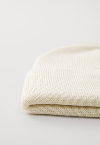 Căciulă tricotată cu nervuri, de culoare crem, cu un tiv pliat, așezată pe o suprafață albă simplă.