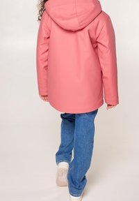 Rosa vattentät jacka med huva och en slät textur, med en något längre bakre nederkant, ihop med blå denimbyxor och vita sneakers.