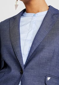 Tmavomodrý sakou s hladkým povrchem, dvěma předními kapsami, jednoduchým zapínáním na knoflík a srdcovým akcentem na kapse. Světle modrá košile pod ním.