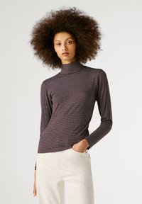 Kõrglaineliste lokkis juustega naine, kes kannab triibulist kõrge kaelusega särki ja heledat pükste kombinatsiooni, poseerib ühe käega taskus.