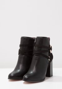 Botas negras hasta el tobillo con un acabado de cuero suave, que cuentan con tiras de gamuza entrelazadas y un acento de hardware en tono dorado. Diseño de tacón alto y grueso.