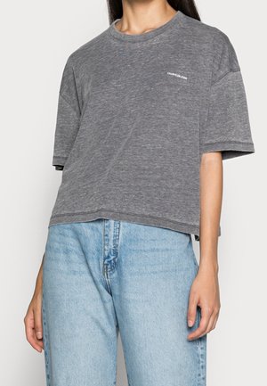 Kobieta ubrana w luźną szarą koszulkę Calvin Klein Jeans i jasnoniebieskie dżinsy z wysokim stanem, stojąca z opuszczonymi ramionami.