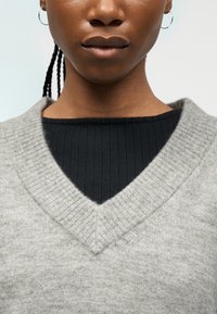 Κοντινό πλάνο ενός ατόμου που φοράει γκρι πουλόβερ V-λαιμόκοψη πάνω από μια μαύρη ριμπ μπλούζα, με πλεγμένα μαλλιά και εμφανές σκουλαρίκι κρίκο.