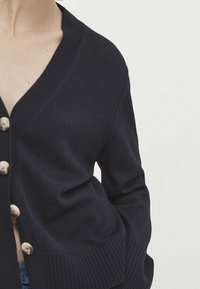 Granatowy sweter o splotach z głębokim dekoltem w kształcie V, z guzikami z przodu, ściągaczami na mankietach i bocznymi kieszeniami, charakteryzujący się miękką teksturą i luźnym krojem.