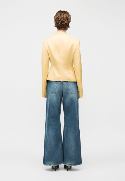 Άτομο με κοντά, σγουρά μαλλιά που φοράει εφαρμοστό κίτρινο μπουφάν, φαρδιά μπλε τζιν και μαύρες ψηλοτάκουνες γόβες, στέκεται με την πλάτη γυρισμένη.