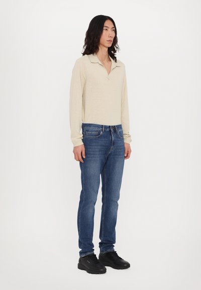 Polo beige à manches longues avec un col, associé à un jean slim bleu foncé et des baskets noires. Tissu lisse sans motifs visibles.