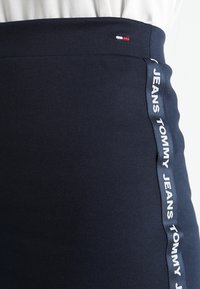 Navyblauwe rok van rekbaar materiaal met een hoge tailleband. Voorzien van een zijstreep met de tekst "TOMMY JEANS" in witte letters.