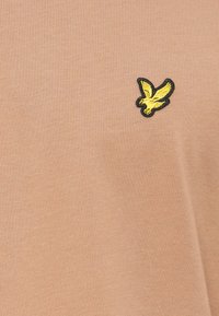 T-shirt en coton beige avec un col rond, présentant un logo d'oiseau brodé noir et jaune sur le côté gauche de la poitrine.