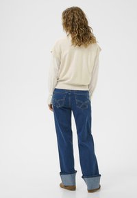 Könnyű bézs top, puffos ujjal, sötétkék farmerral párosítva, amelynek felgöngyölt szárai és részletes hátsó zsebei vannak.