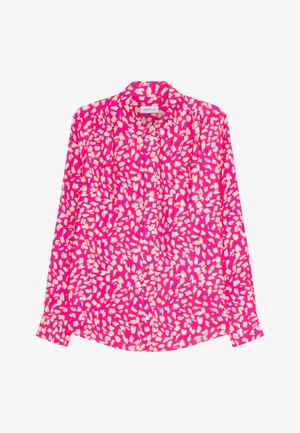 Persoon draagt een roze overhemd met lange mouwen en witte abstracte vlekken, en lichtbeige broek, handen in de zakken.