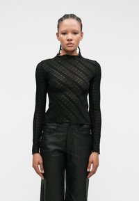 Svart langermet topp laget av teksturert blonde, med bølgete stripemønster og høy hals. Kombinert med høyhalsede svarte bukser.