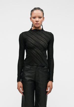 Czarny top z długim rękawem wykonany z teksturowanej koronki, z falistym wzorem w paski i wysokim dekoltem. Połączony z czarnymi spodniami o wysokim stanie.