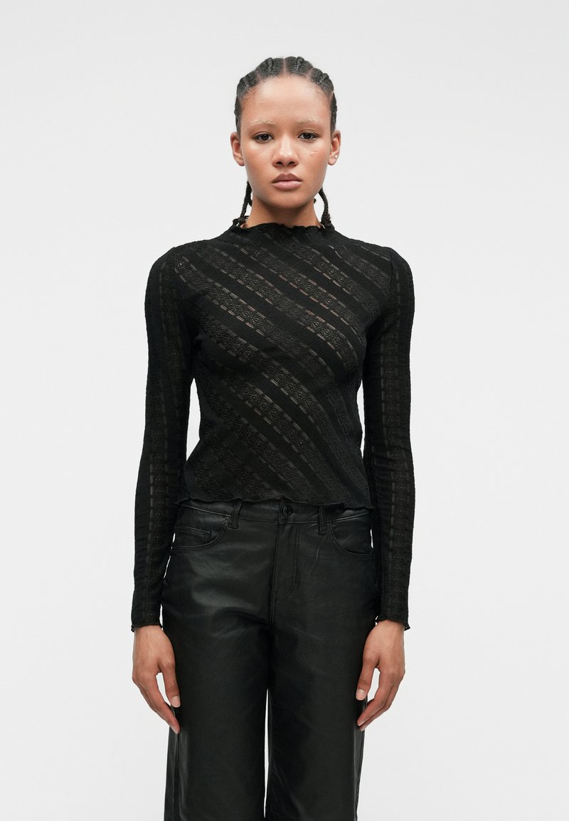 Svart langermet topp laget av teksturert blonde, med bølgete stripemønster og høy hals. Kombinert med høyhalsede svarte bukser.