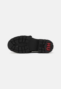Semelle de basket noire avec des profondeurs de semelle pour traction, dotée d'une texture en caoutchouc et du logo rouge "HU" sur le talon extérieur.