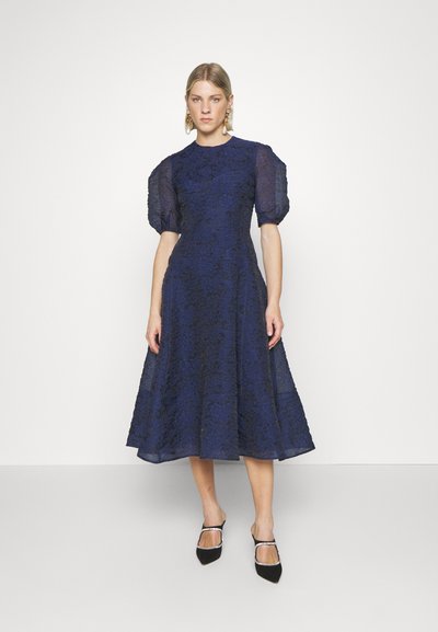 Vestido azul marinho com mangas curtas bufantes, textura de renda floral e silhueta A-line. Combinado com sapatos pretos de bico fino.