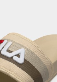 Μπεζ σανδάλι χωρίς λουράκια με υφασμάτινο λουράκι με ανάγλυφη υφή, διακοσμημένο με ανάγλυφα λευκά και κόκκινα κεντημένα γράμματα.