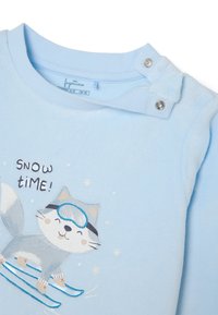 Haut bleu clair à manches longues avec fermeture à pression. Présente une illustration d'un chat skiant, accompagnée du texte "TEMPS DE NEIGE !" en marron foncé.