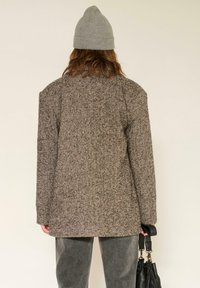 Blazer oversize marron en mélange de laine à motif chevrons, avec épaules rembourrées, larges revers et coupe droite, associé à un pantalon gris et un bonnet.