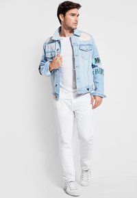 Heleheline teksajakk, millel on pleekinud laigud, abstraktsed mustrid ja must kiri varrukatel, kantud üle valge T-särgi ja valgete teksade.