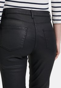 Schwarze, dehnbare Jeans aus Denim mit glatter Textur, ausgestattet mit zwei Gesäßtaschen und Gürtelschlaufen, seitlich präsentiert mit einem teilweise sichtbaren gestreiften Oberteil.