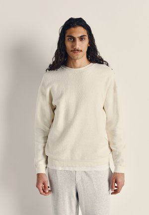 Cream-colored fleece sweatshirt with a textured surface, round neck, and ribbed cuffs. Paired with light gray sweatpants.