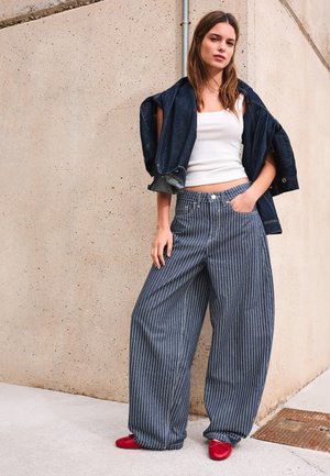 Žena stojí oproti béžovej stene, má na sebe biele tielko, tmavú rifľovú bundu prehodenú cez ramená, modré široké nohavice s prúžkami a červené topánky.