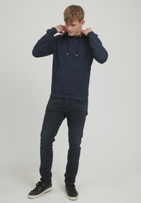 Marineblå hettegenser med kengurulomme, snøring og ribbestrikket mansjett, kombinert med mørke jeans og svarte joggesko.