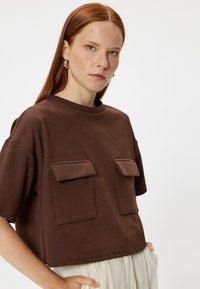 Brązowy krótki t-shirt z krótkimi rękawami, wyposażony w dwie kieszenie z przodu. Wykonany z miękkiego materiału, ma okrągły dekolt i luźny krój.