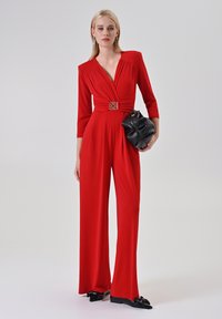 Donna che indossa una tuta rossa a maniche lunghe con una cintura decorativa, tiene una pochette nera testurizzata e indossa delle ballerine nere a punta con fiocchi.