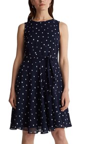 Femme portant une robe bleu marine sans manches à pois blancs et une ceinture nouée à la taille, debout, les bras détendus.