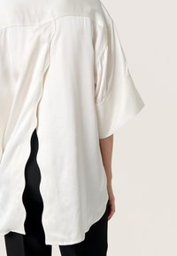 Blusa crema ampia realizzata in tessuto morbido, con orlo a scallop e maniche corte, abbinata a pantaloni neri.