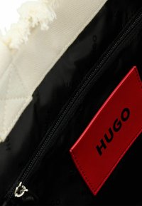 Interno di borsa in nylon nero con etichetta in pelle rossa con il marchio "HUGO" in rilievo e tessuto crema strutturato. Chiusura con zip e dettagli cuciti.