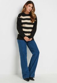 Maglione a righe nero e crema con maniche lunghe, texture a costine e dettagli con bottoni sulle spalle, abbinato a jeans blu a zampa d'elefante.