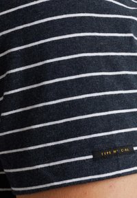 Chemise à manches courtes bleu marine avec des rayures horizontales blanches, présentant une texture douce et une étiquette noire sur l'ourlet avec un texte blanc.