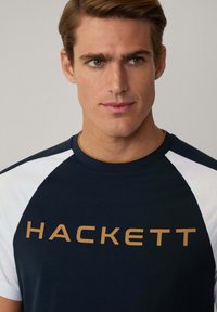 Navy modrá tričko s bielymi raglánovými rukávmi, s veľkým zlatým logom "HACKETT". Zmes bavlny s jemnou textúrou.