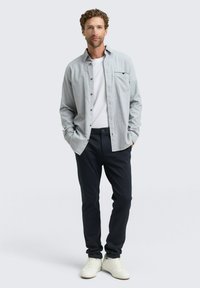 Uomo con capelli ricci che indossa una camicia grigia chiara a bottoni sopra una maglietta bianca, pantaloni blu scuro e sneakers bianche, in piedi con le mani nelle tasche.