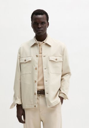 Man draagt licht beige blouse met knopen, bijpassende broek, beige poloshirt en donkere riem, poseert tegen een effen witte achtergrond.