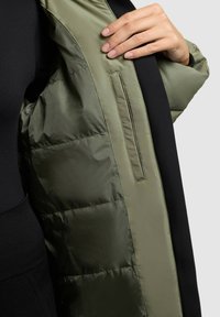 Gevoerde jas in olijfgroen met een gladde afwerking en een zwarte stof aan de buitenkant. Voorzien van een zijzak en een doorgestikt patroon.