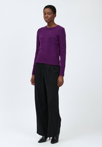 Frau mit einem strukturierten lila Pullover, weiten schwarzen Hosen und schwarzen spitzen Stiefeletten mit Absatz, die vor einem schlichten weißen Hintergrund steht.