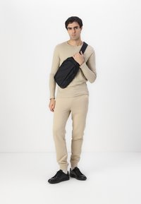 Svart crossover-veske laget av stoff, med flere glidelåslommer og et slankt design. Modellen har på seg en beige langermet topp og bukser.