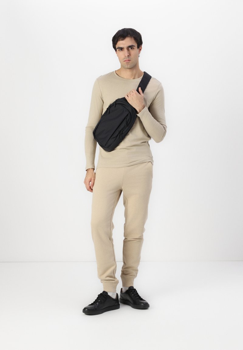 Svart crossover-veske laget av stoff, med flere glidelåslommer og et slankt design. Modellen har på seg en beige langermet topp og bukser.