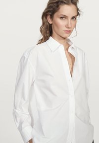 Camicia bianca con bottone, caratterizzata da un colletto classico, maniche lunghe e una vestibilità rilassata. Il tessuto appare morbido e leggero con dettagli minimi.