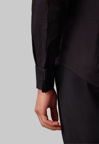 Bras droit d'une personne portant une chemise noire à manches longues avec poignet boutonné et un pantalon noir sur un fond gris.