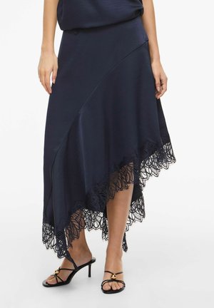 Naine, kes kannab tumesinist asümmeetrist seelikut pitsilise äärisega, mida täiendavad mustad rihmikud kõrge kontsaga sandaalid.